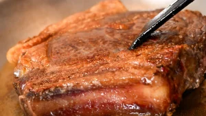 Tongs touching a golden-brown chuleton steak as it sears on its second side in a pan, Spain.