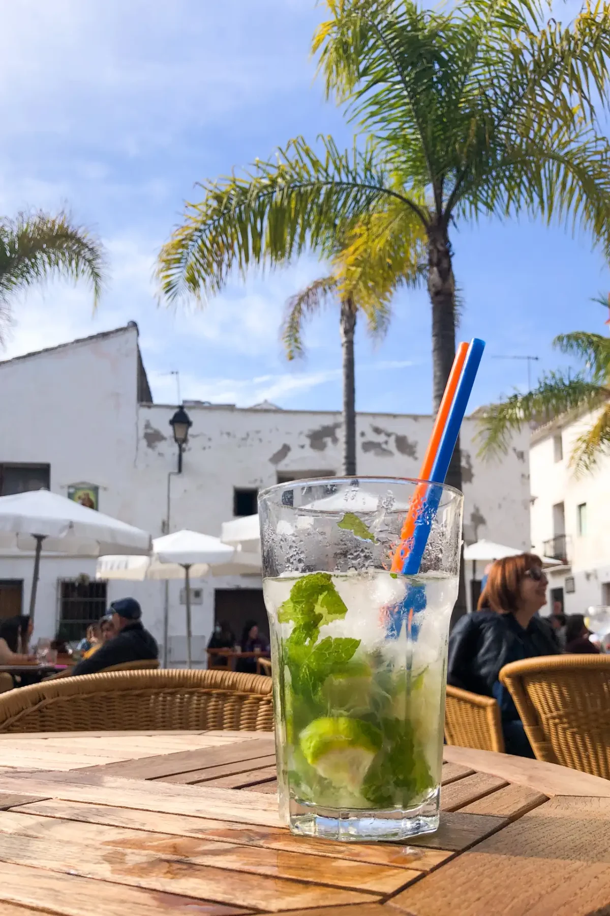 A mojito with mint, lime and ice on a wooden terrace table, palm trees and white buildings in a sunny plaza, Dénia, Spain