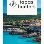 Tapas Hunters Issue 03 cover — Llanes harbour with turquoise water, rocky coastline and orange-roofed town, Asturias, Spain