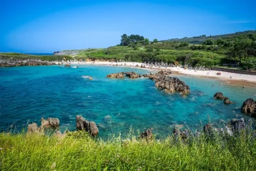 Llanes coastline — turquoise rocky bay with a wide sandy beach, jagged rock formations and wildflowers in the foreground, Asturias