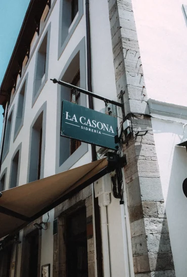 Sidrería La Casona hanging sign on a stone corner building with a tan awning below, Llanes, Asturias