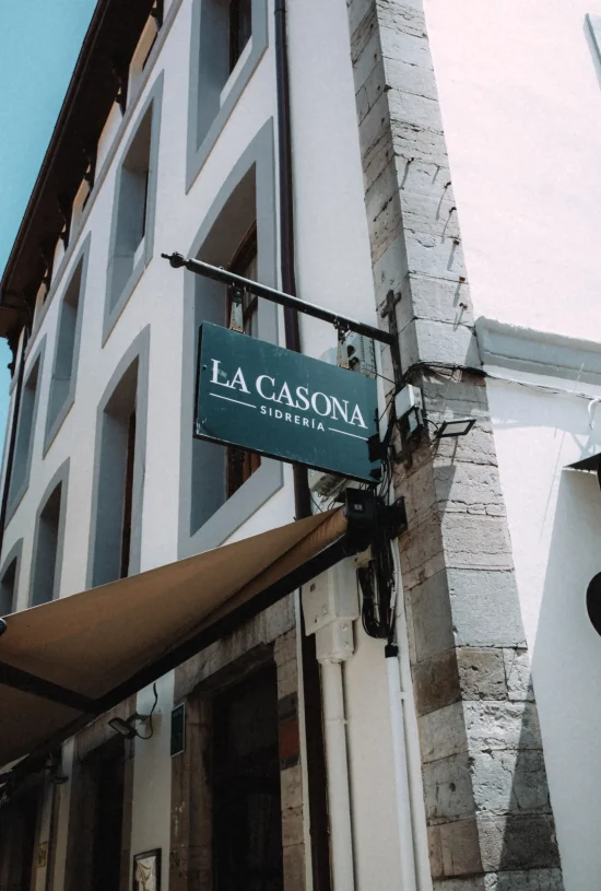 Sidrería La Casona hanging sign on a stone corner building with a tan awning below, Llanes, Asturias