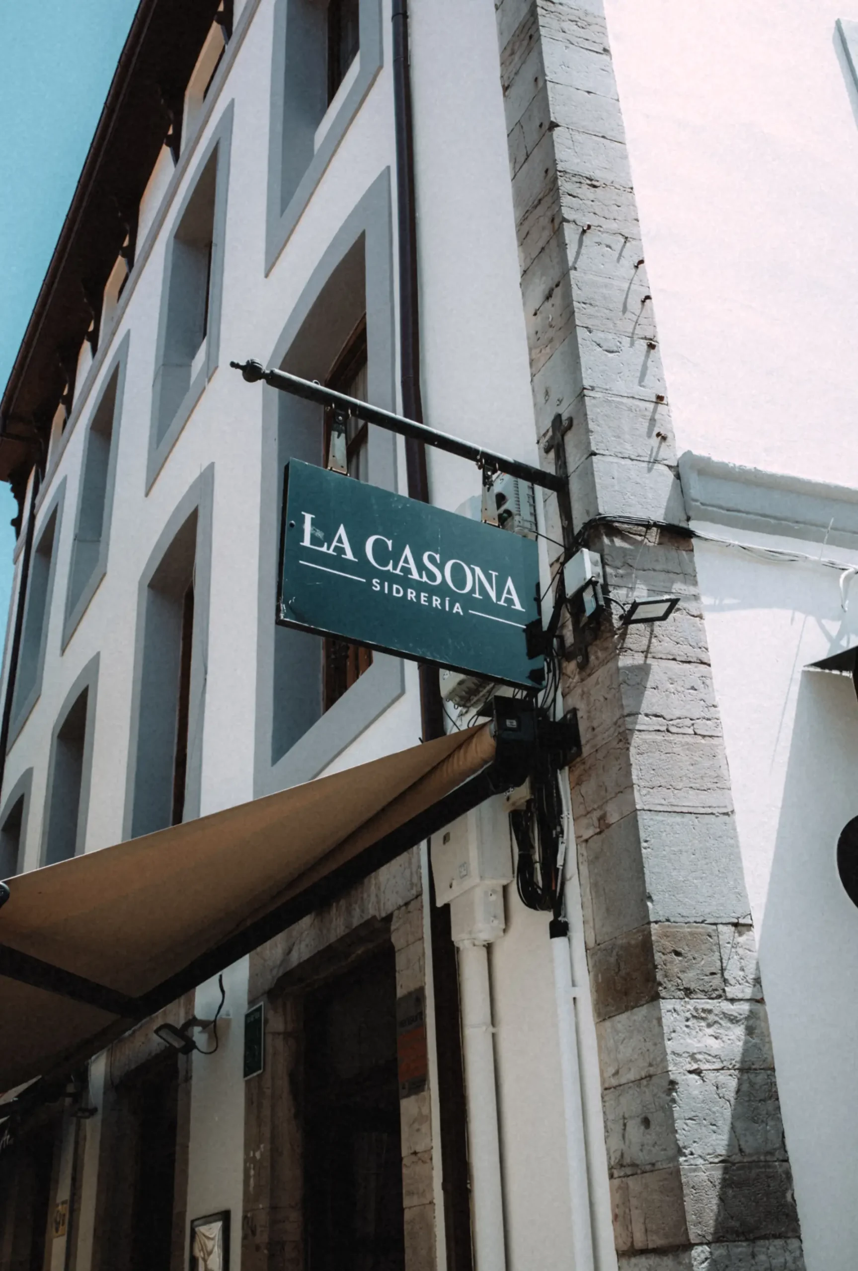 Sidrería La Casona hanging sign on a stone corner building with a tan awning below, Llanes, Asturias