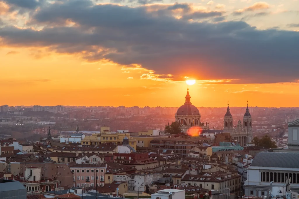 Madrid skyline at sunset — Almudena Cathedral dome and spire silhouetted against a fiery orange sky, rooftops stretching to the horizon