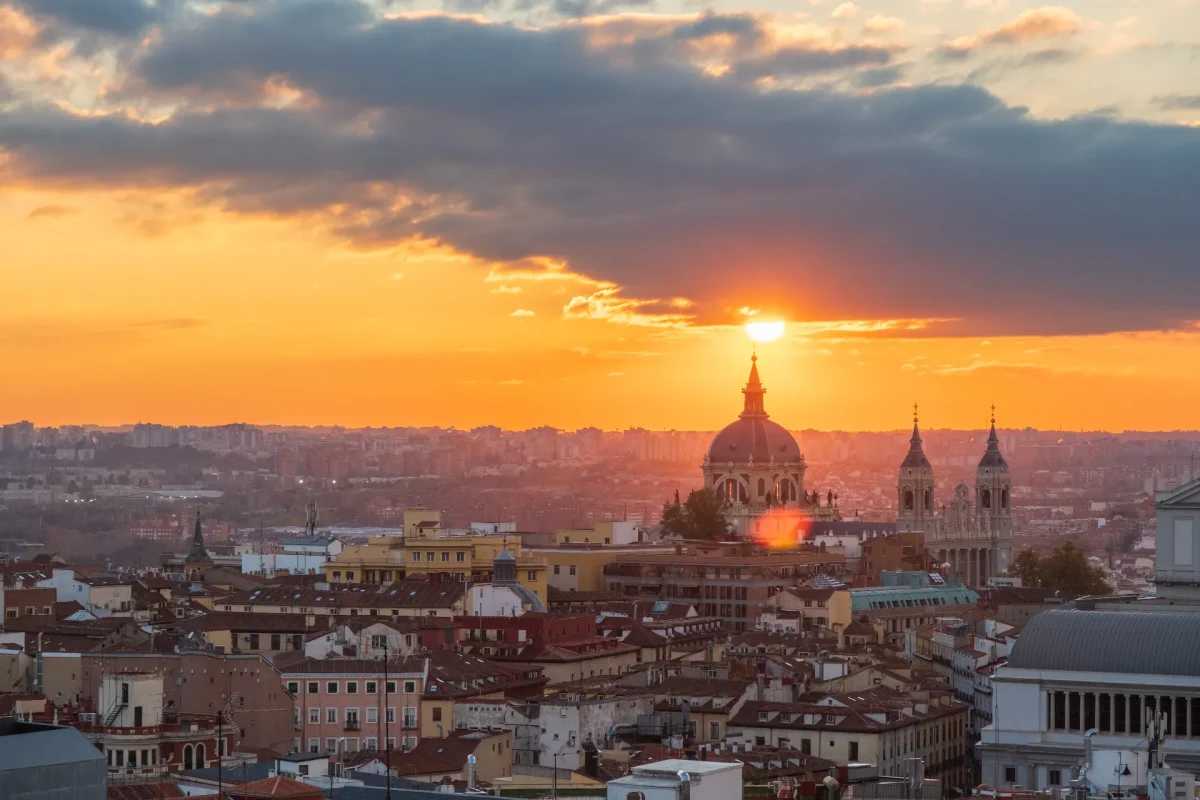 Madrid skyline at sunset — Almudena Cathedral dome and spire silhouetted against a fiery orange sky, rooftops stretching to the horizon