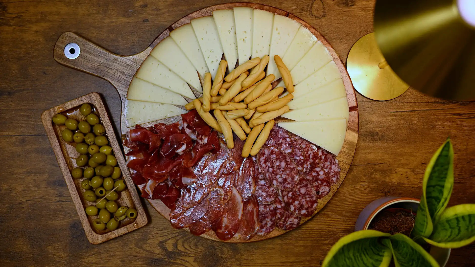 Spanish picoteo board — sliced cheese, jamón, cured meats, breadsticks and green olives on a round wooden board, Spain