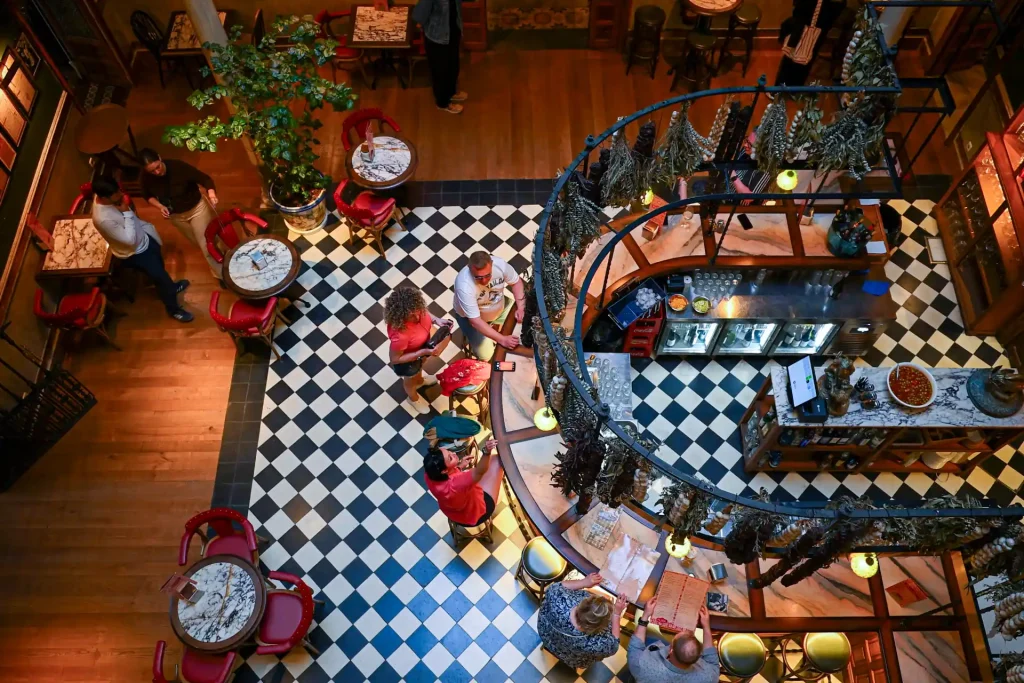 Spanish bar interior from above — black and white diamond floor tiles, red chairs, a curved bar counter and dried herbs hanging overhead
