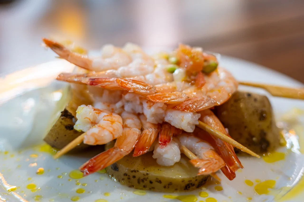 Skewered langoustines layered over sliced potatoes drizzled with oil on a white plate at Chantarela, Spain.