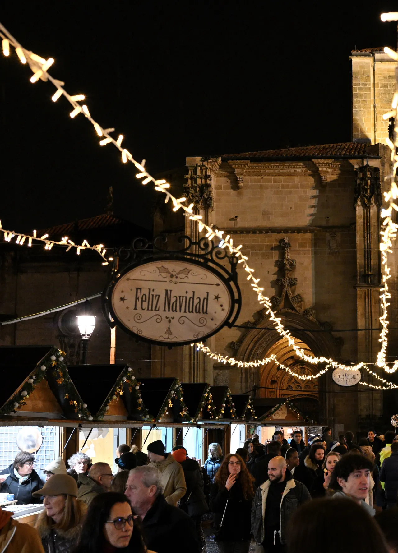 Nighttime view of a festive Christmas market with wooden stalls, string lights, and a 