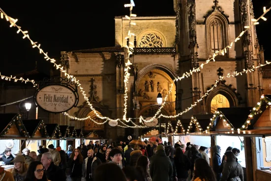 Nighttime view of a bustling Christmas market with wooden stalls, string lights, and holiday crowds set against the historic facade of the Oviedo Cathedral in Spain.