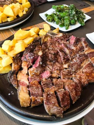A sliced chuleton steak with fried potatoes and a side of padron peppers on a dark platter at El Papatorio, Santiago de Compostela.