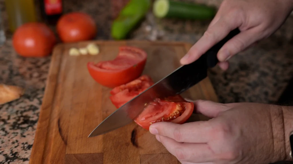 Close-up of hands using a knife to slice ripe red tomatoes on a wooden cutting board, with other ingredients like garlic and cucumber in the background.