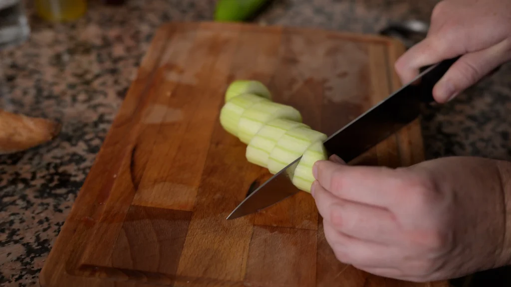 Close-up of hands slicing a peeled cucumber on a wooden cutting board with a kitchen knife.