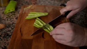 Close-up of hands using a knife to chop green peppers on a wooden cutting board.