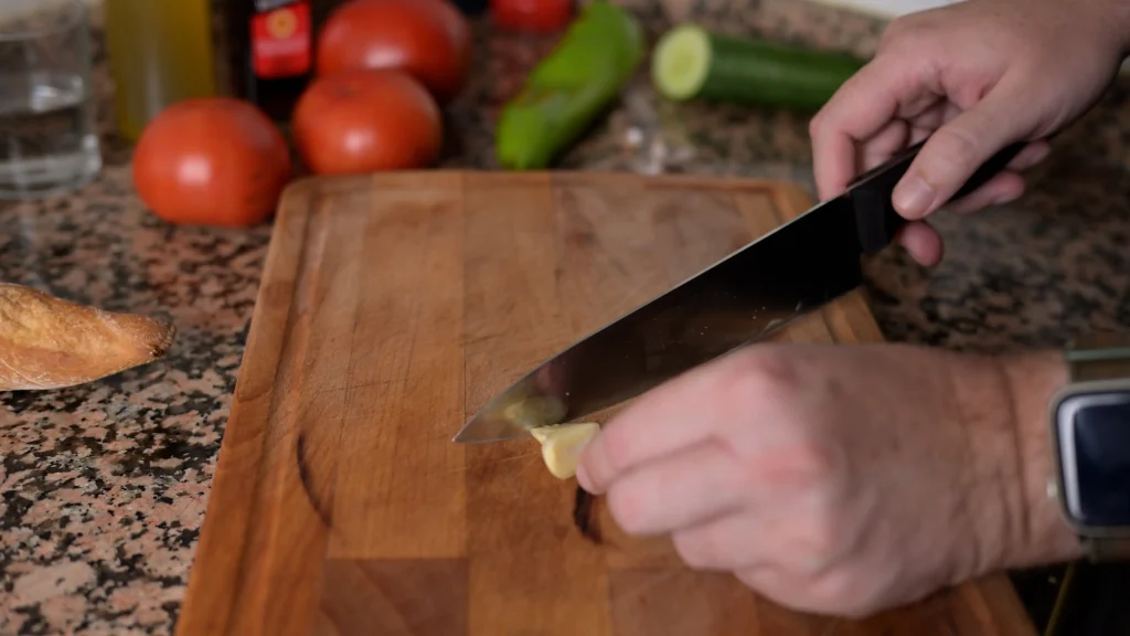Close-up of hands using a knife to chop garlic cloves on a wooden cutting board.