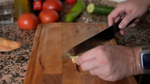 Close-up of hands using a knife to chop garlic cloves on a wooden cutting board.