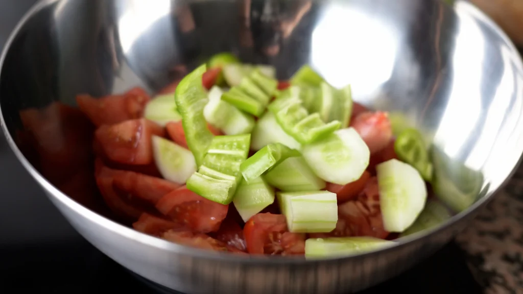 Close-up of a stainless steel mixing bowl filled with chopped fresh tomatoes, sliced cucumbers, and green peppers for making gazpacho.