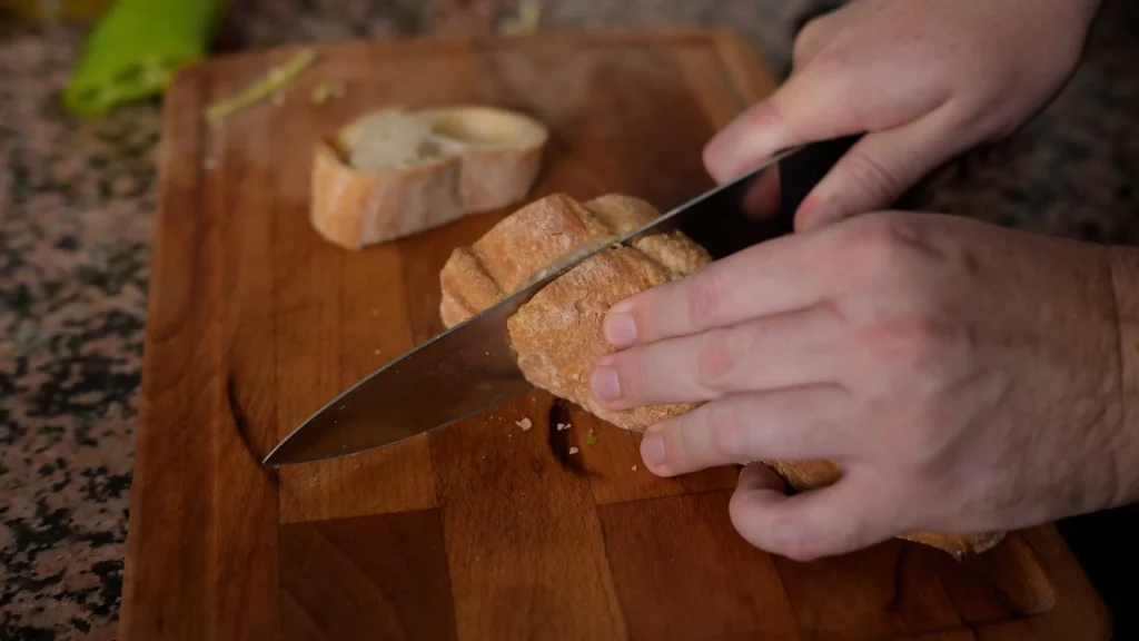 Close-up of hands using a knife to slice crusty bread on a wooden cutting board.