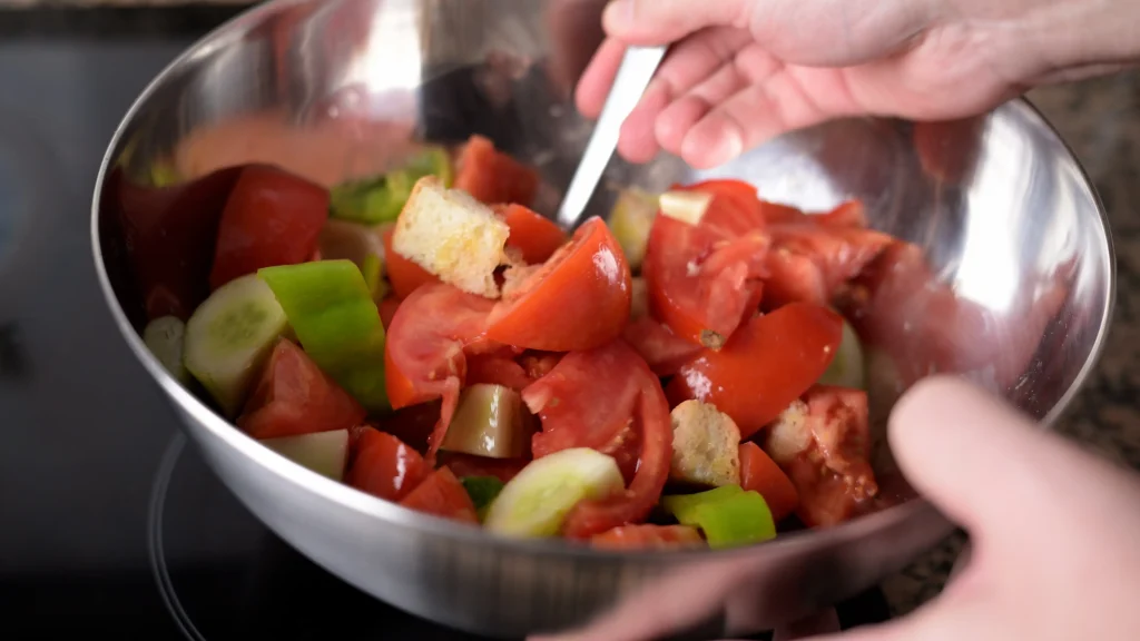Close-up of hands mixing chopped tomatoes, cucumbers, green peppers, and bread in a stainless steel bowl with a utensil.