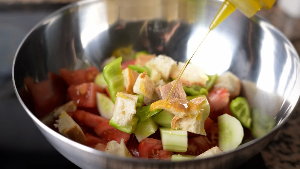 Close-up of olive oil being poured from a bottle onto chopped tomatoes, cucumbers, green peppers, and bread in a stainless steel mixing bowl.