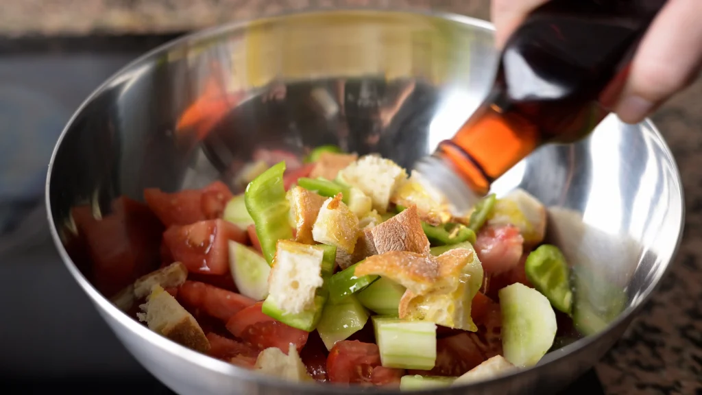 Close-up of vinegar being poured from a bottle onto chopped vegetables and bread in a stainless steel mixing bowl.