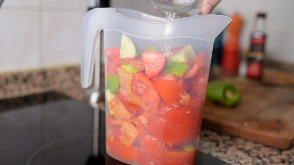 Close-up of water being poured from a glass into a large plastic pitcher filled with chopped tomatoes, cucumbers, green peppers, and bread.