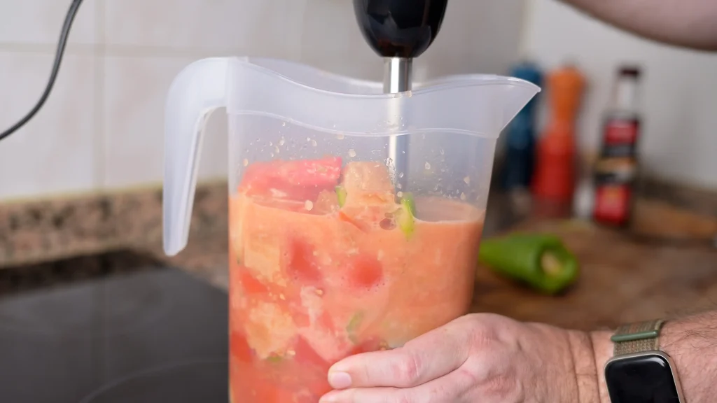 Close-up of an immersion blender being used to puree chopped vegetables and bread inside a large plastic pitcher for gazpacho andaluz.