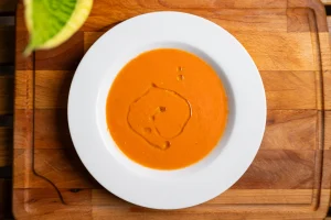 Overhead view of a vibrant red bowl of gazpacho soup garnished with a drizzle of olive oil, resting on a white plate over a wooden cutting board.