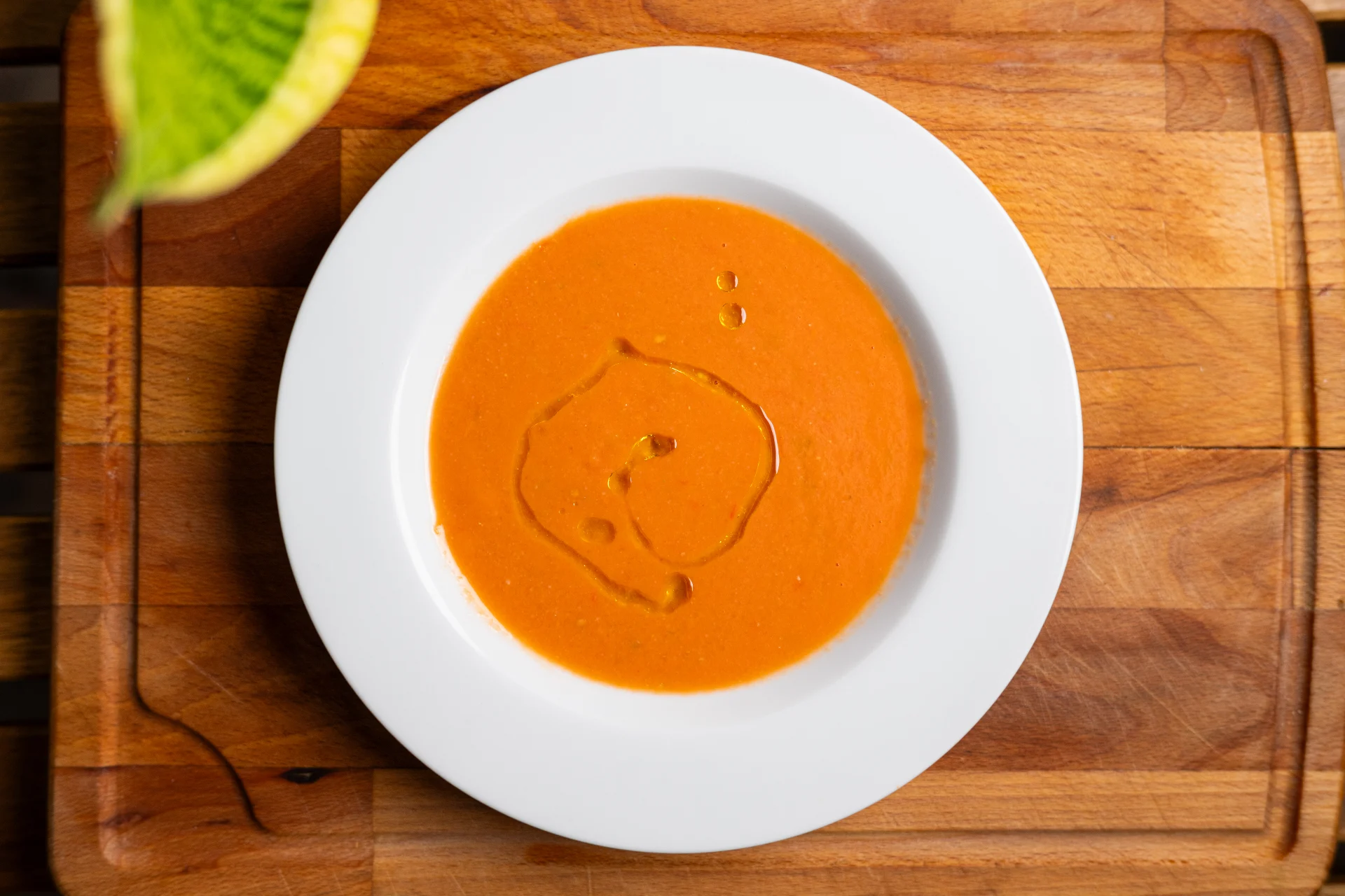 Overhead view of a vibrant red bowl of gazpacho soup garnished with a drizzle of olive oil, resting on a white plate over a wooden cutting board.