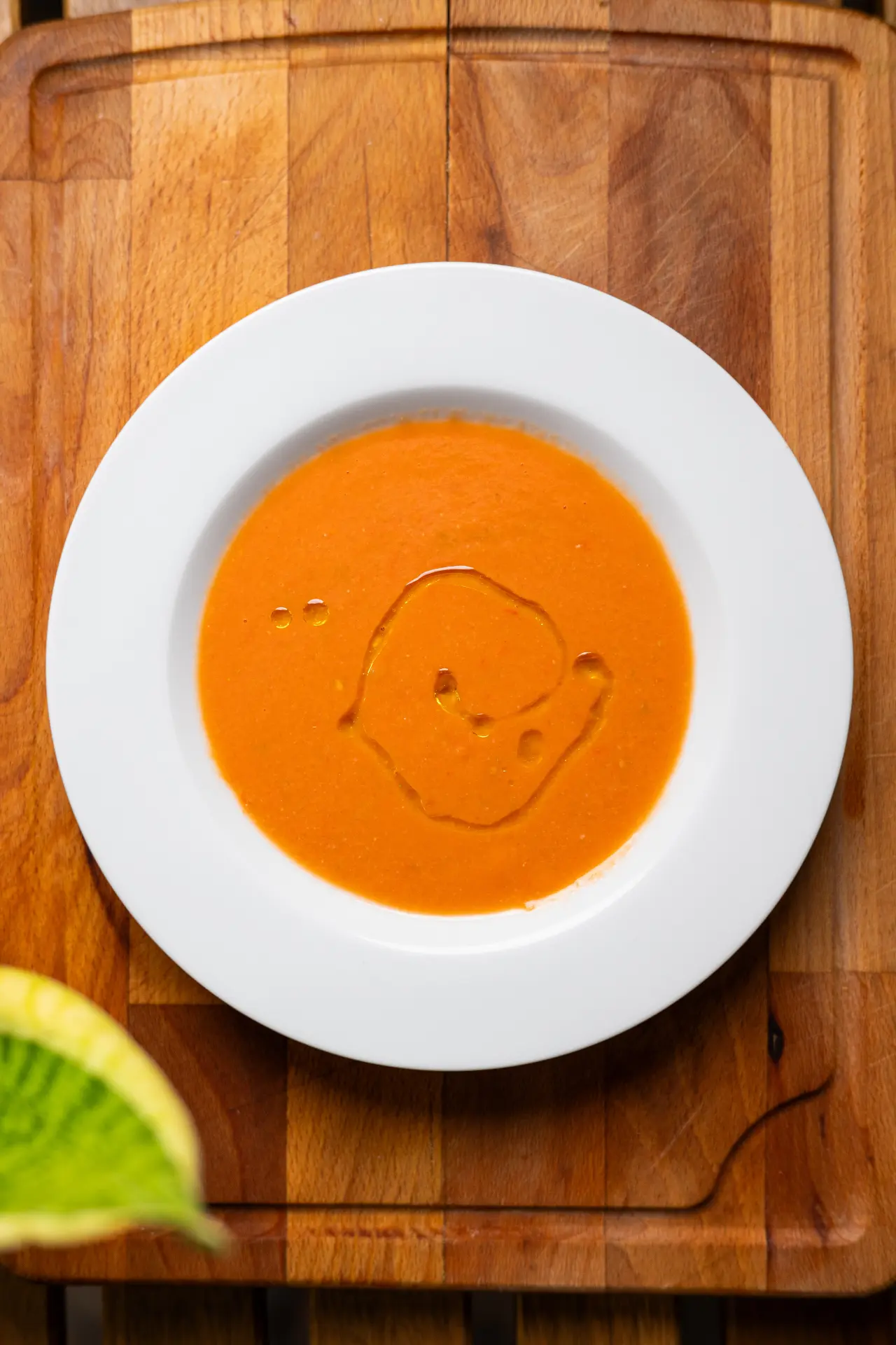 Overhead view of a vibrant red bowl of gazpacho soup garnished with a drizzle of olive oil, resting on a white plate over a wooden cutting board.