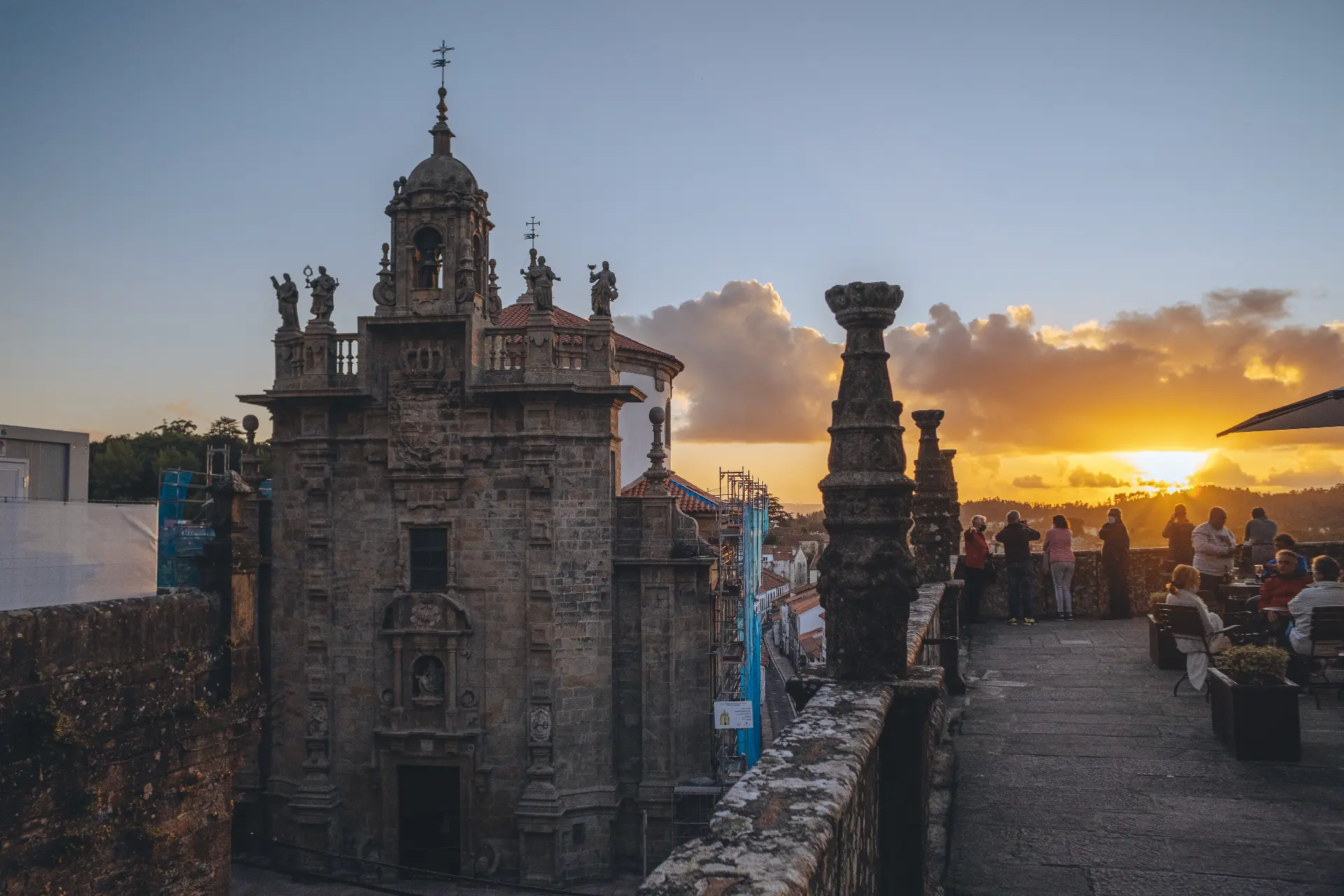 The ornate stone tower of a historic building and people gathered on a rooftop terrace at sunset, Santiago de Compostela.