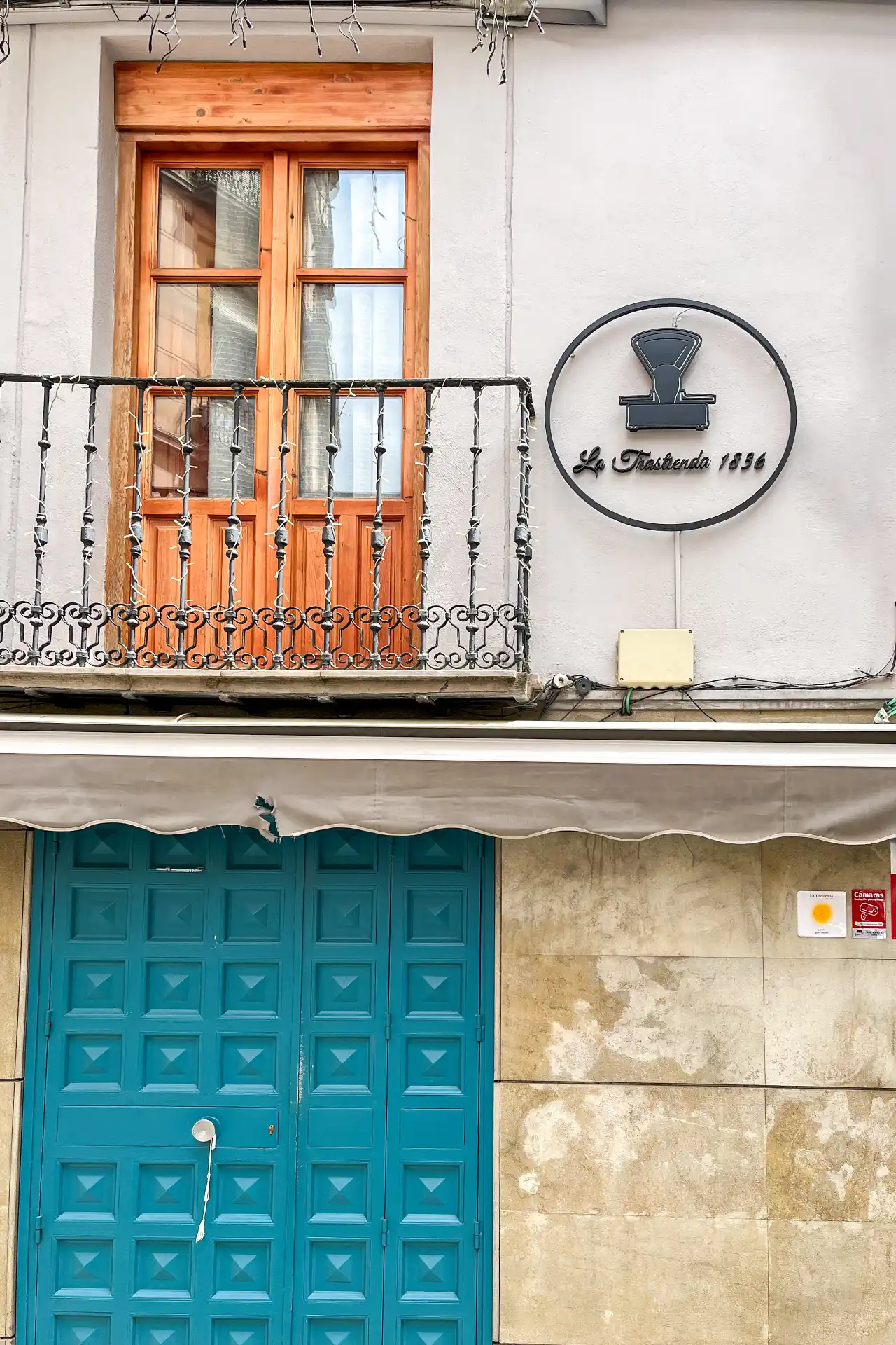 The exterior facade of La Trastienda 1836 featuring a bright teal paneled door, a balcony with wooden shutters, and a circular sign in Granada, Spain.