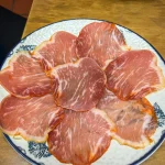Lomo ibérico de bellota - thin-sliced cured pork loin fanned on a blue-rimmed ceramic plate, sliced tomatoes on a second plate behind, La Trastienda, Granada