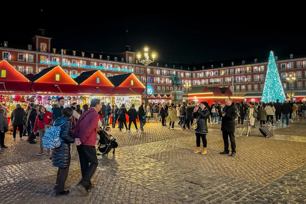 Nighttime view of the bustling Plaza Mayor Christmas market with illuminated red stalls, festive holiday lights, and a glowing Christmas tree in Madrid, Spain.
