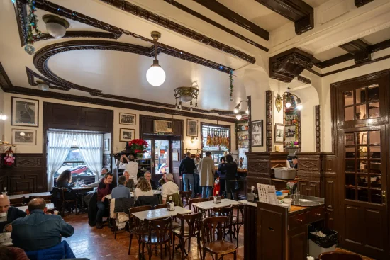 Interior view of a traditional, bustling restaurant or tapas bar with dark wood paneling, ornate ceilings, and patrons seated at tables in Madrid, Spain.