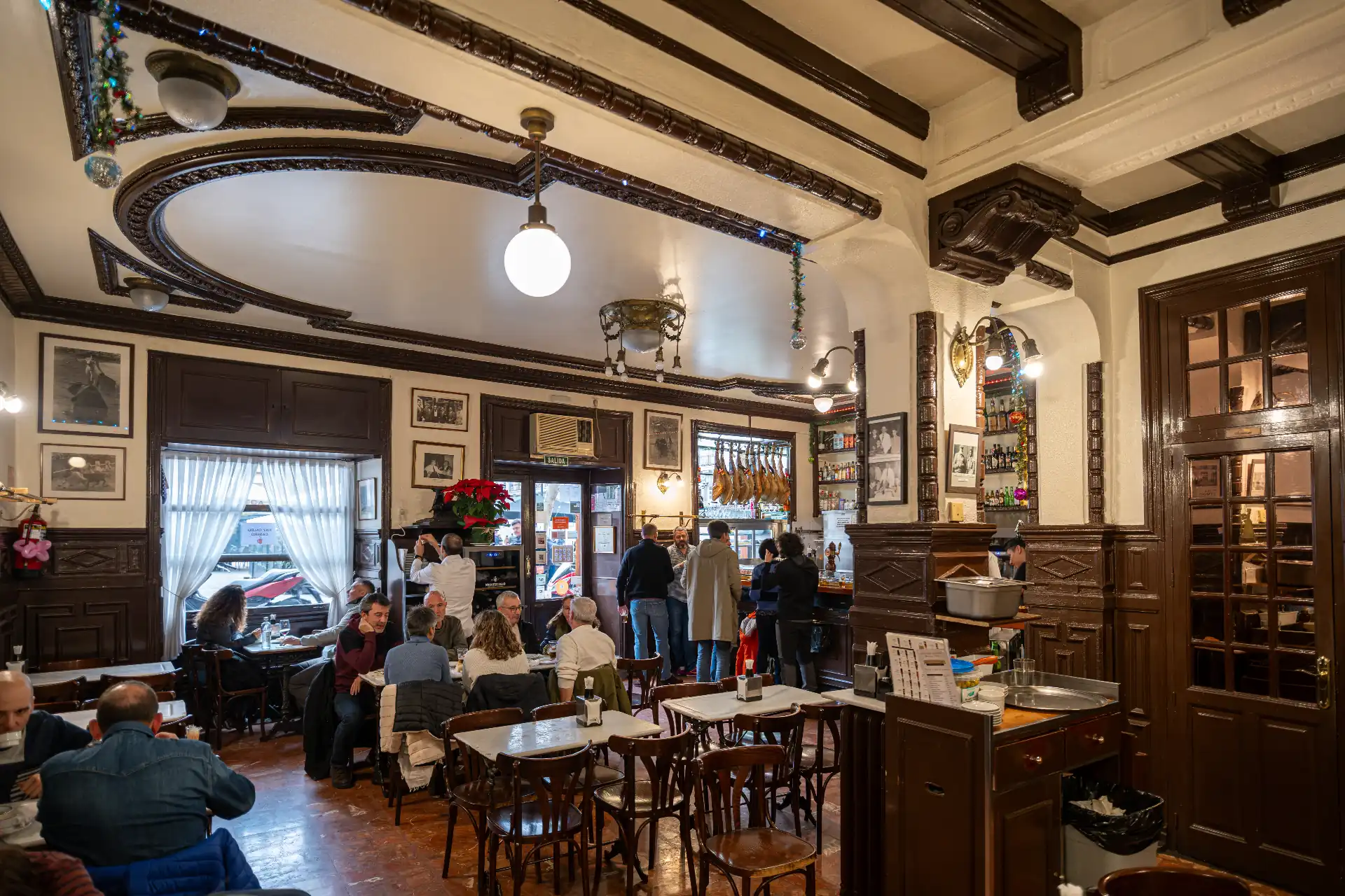 Interior view of a traditional, bustling restaurant or tapas bar with dark wood paneling, ornate ceilings, and patrons seated at tables in Madrid, Spain.