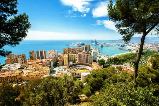Elevated panoramic view of Málaga, Andalusia, Spain, featuring the bullring, cityscape, port, and coast framed by green trees under a bright blue sky.