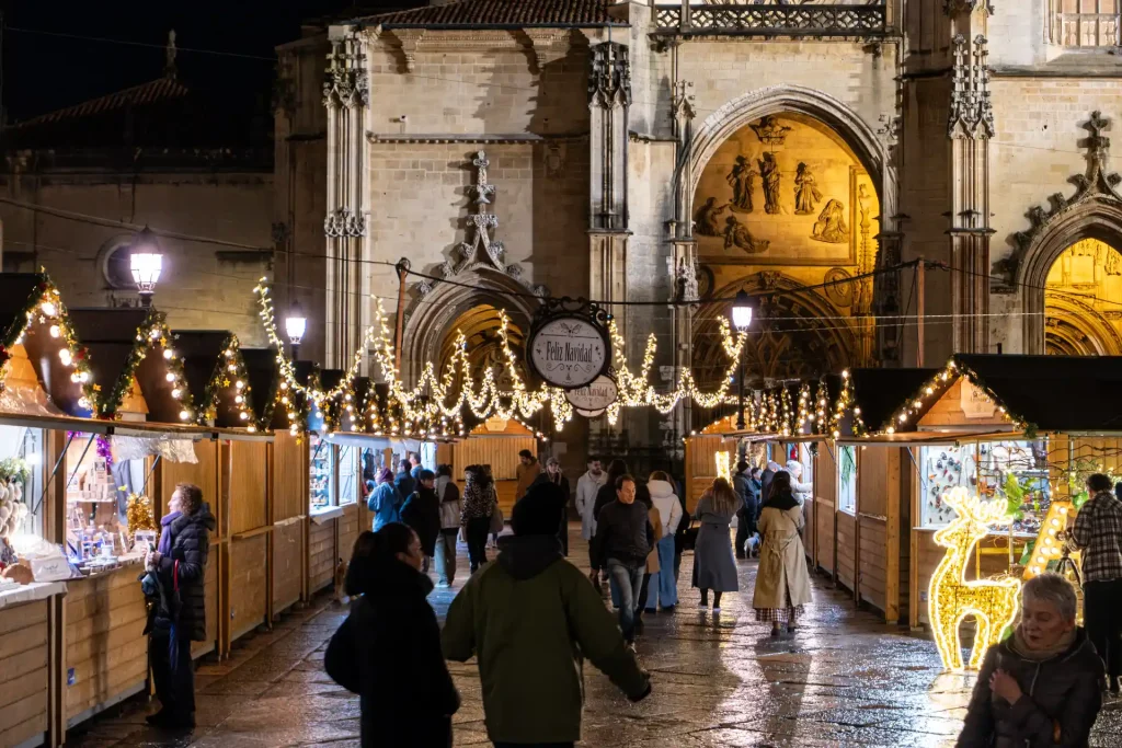Nighttime view of a festive Christmas market with wooden stalls, string lights, and holiday crowds set against the historic facade of the Oviedo Cathedral in Spain.