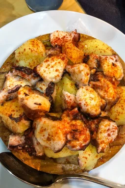 Pulpo a feira — boiled octopus pieces on potato slices, heavily dusted with paprika and olive oil, on a wooden board, El Papatorio