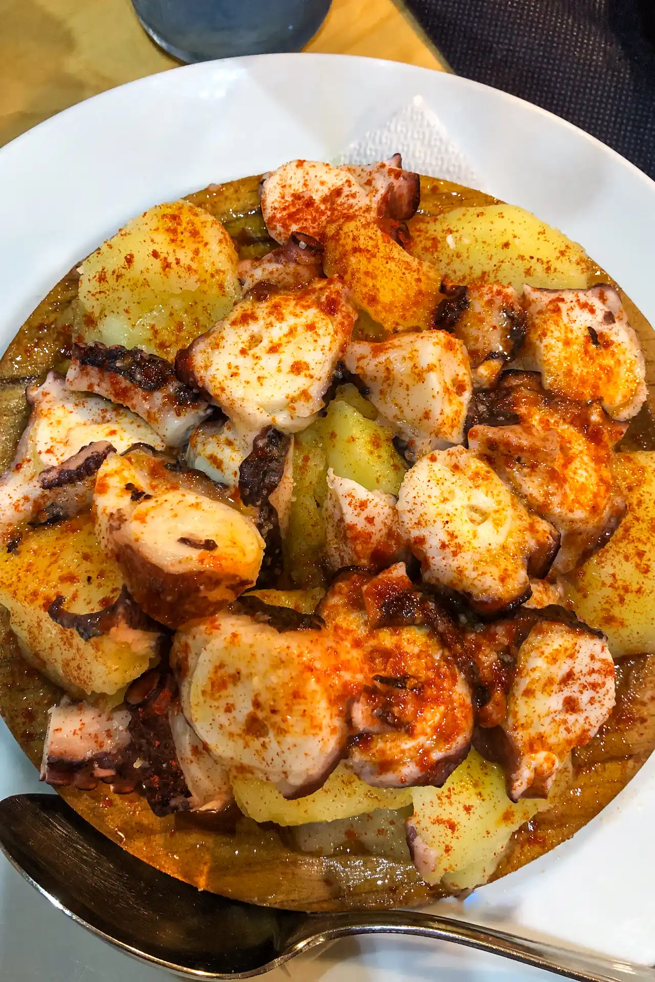 Pulpo a feira — boiled octopus pieces on potato slices, heavily dusted with paprika and olive oil, on a wooden board, El Papatorio