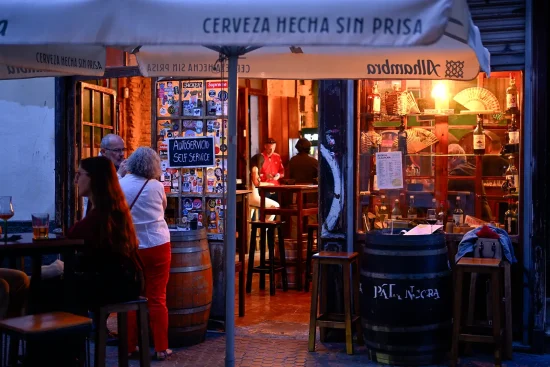 Exterior view of a cozy traditional tapas bar with outdoor barrel tables, patrons sitting and standing under umbrellas, and warm interior lighting in Spain.