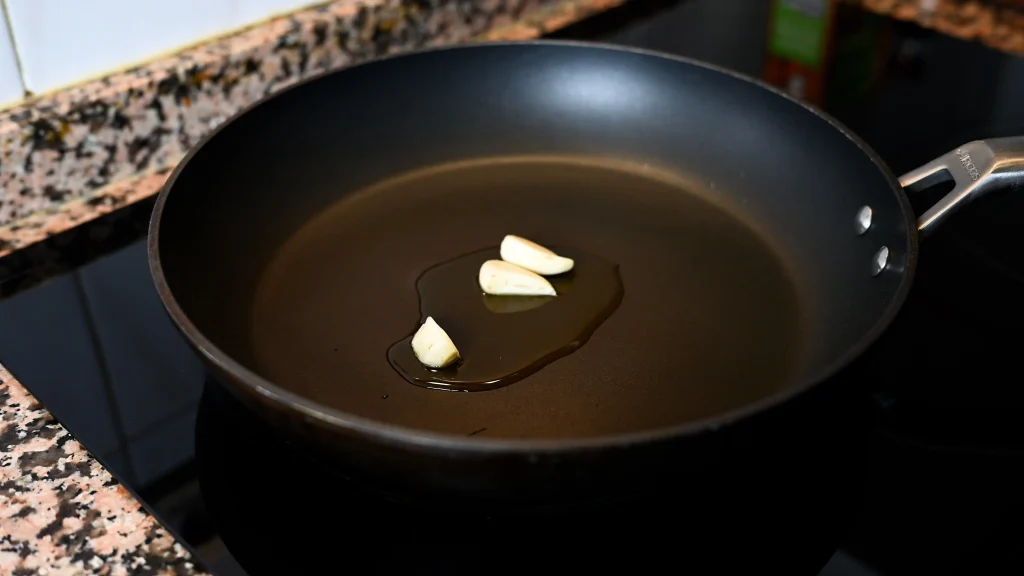 Close-up of garlic cloves frying in olive oil in a black pan on a stovetop during step 2 of the garbanzos con espinacas recipe for Tapas Hunters