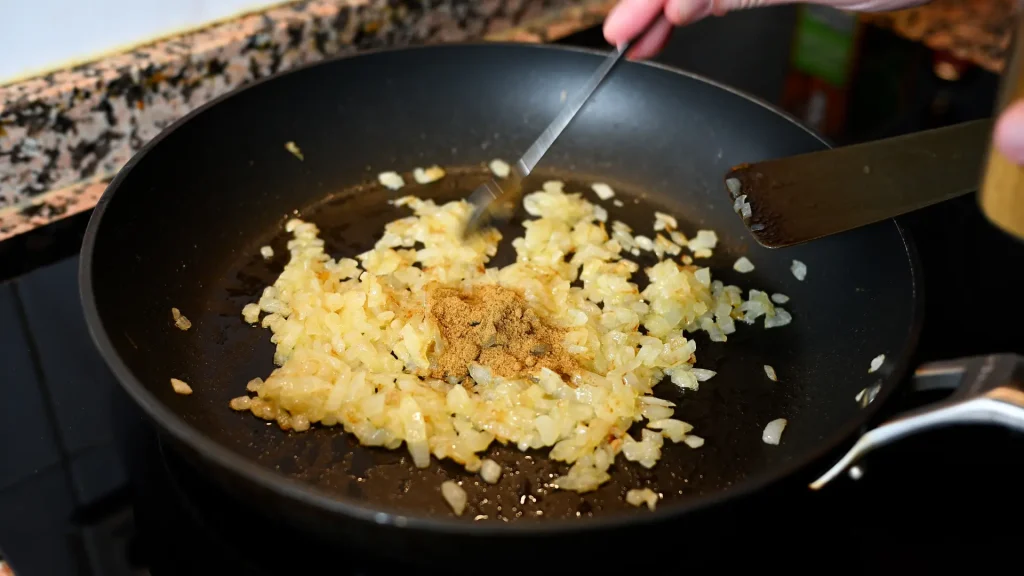 Close-up of adding ground cumin spice to sautéed onions in a pan during step 8 of the garbanzos con espinacas recipe for Tapas Hunters.