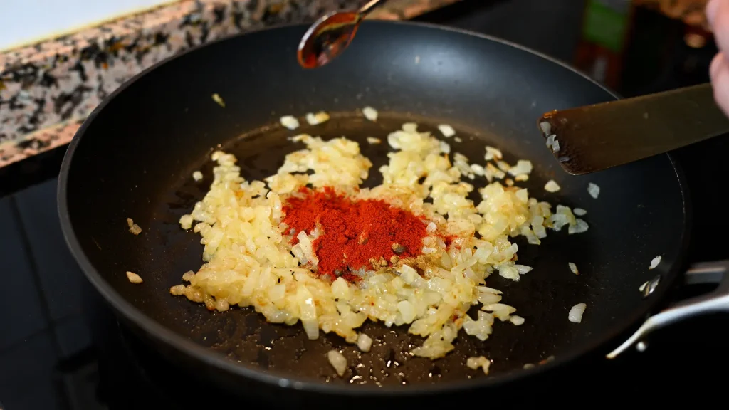 Close-up of adding Spanish paprika to sautéed onions in a pan during step 9 of the garbanzos con espinacas recipe for Tapas Hunters.