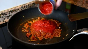 Close-up of pouring canned crushed tomatoes into a pan with sautéed onions and spices during step 10 of the garbanzos con espinacas recipe for Tapas Hunters.