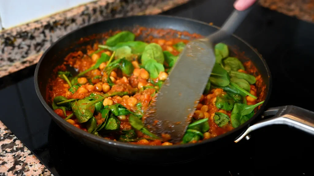 Close-up of folding fresh spinach leaves and chickpeas into the rich simmering sauce with a spatula during step 15 of the garbanzos con espinacas recipe for Tapas Hunters.