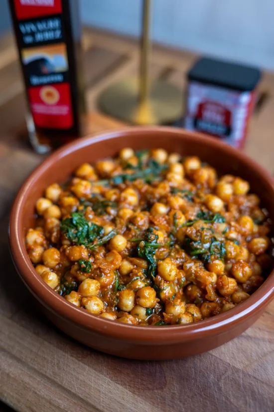 Close-up of a traditional Spanish chickpea and spinach stew (garbanzos con espinacas) served in a terracotta bowl with bottles of sherry vinegar and spices in the background.