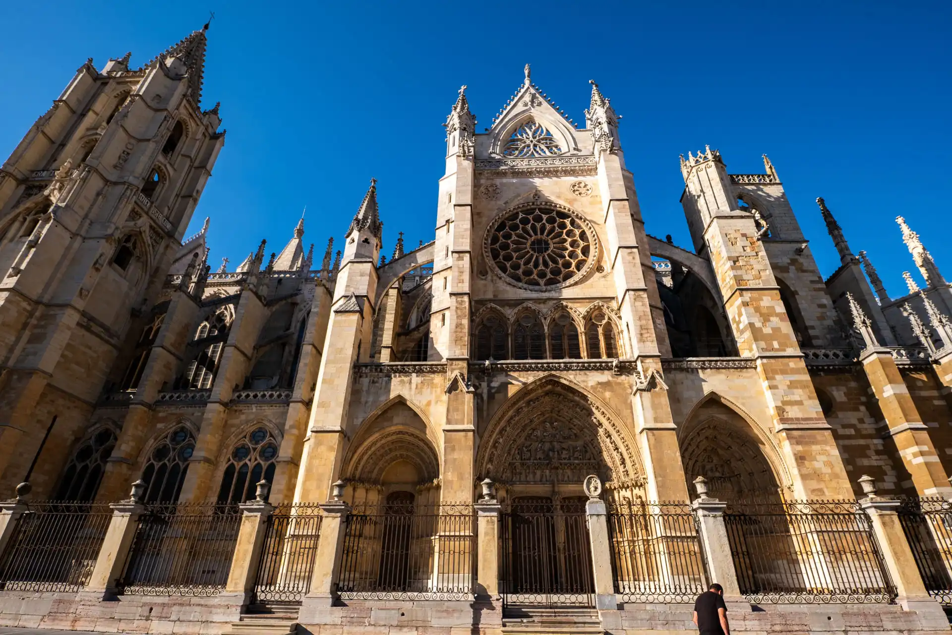 Low-angle view of the ornate Gothic facade, towers, and intricate rose window of the León Cathedral under a clear blue sky in Spain.