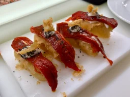 Close-up of four gourmet tapas featuring crispy fried bases topped with roasted red peppers, a dollop of white sauce, and black seasoning flakes on a white board.