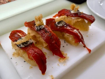 Close-up of four gourmet tapas featuring crispy fried bases topped with roasted red peppers, a dollop of white sauce, and black seasoning flakes on a white board.
